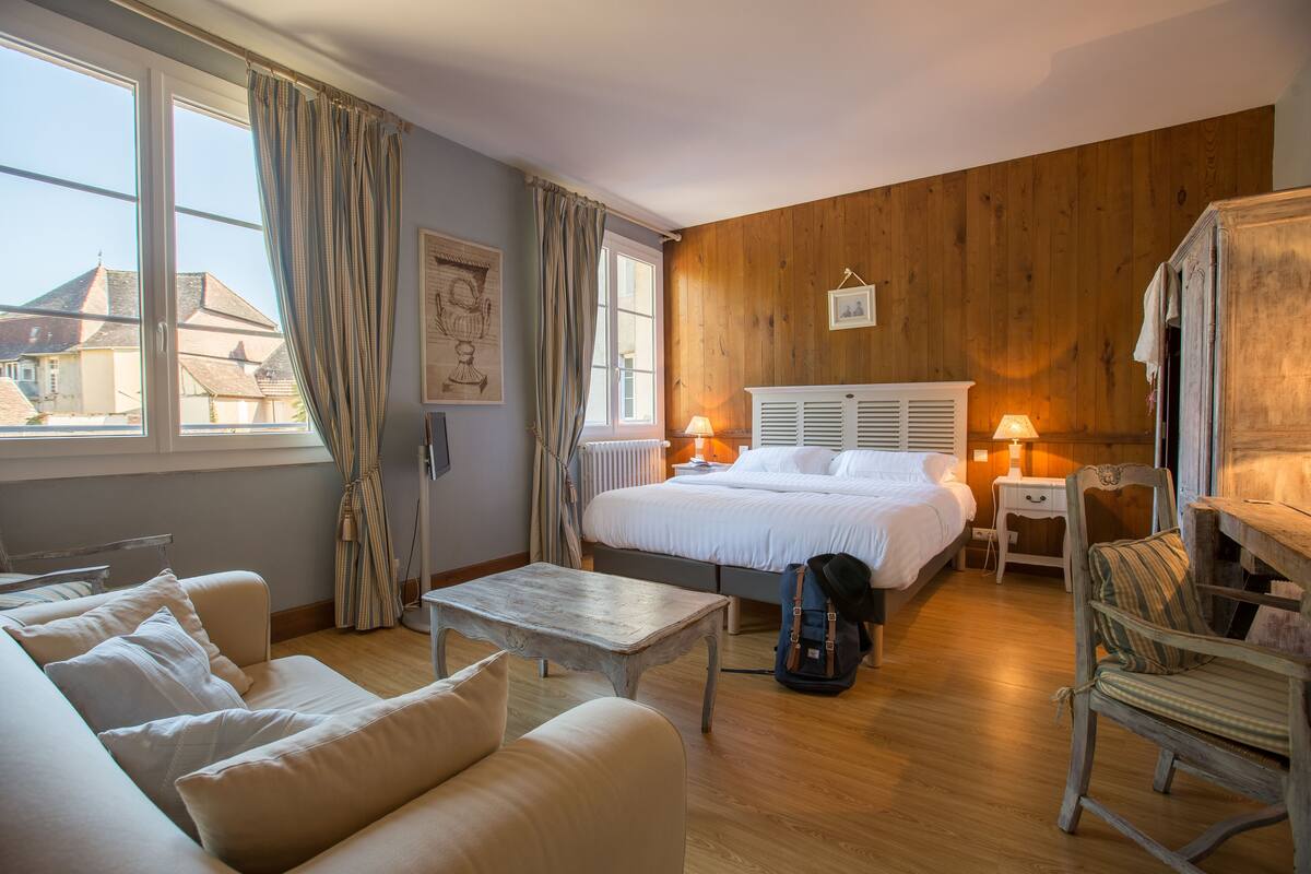 Ein Schlafzimmer mit einem großen Bett, Nachttischen mit Lampen, einem hölzernen Kopfteil, einem Fenster mit Vorhängen und Blick auf Gebäude.