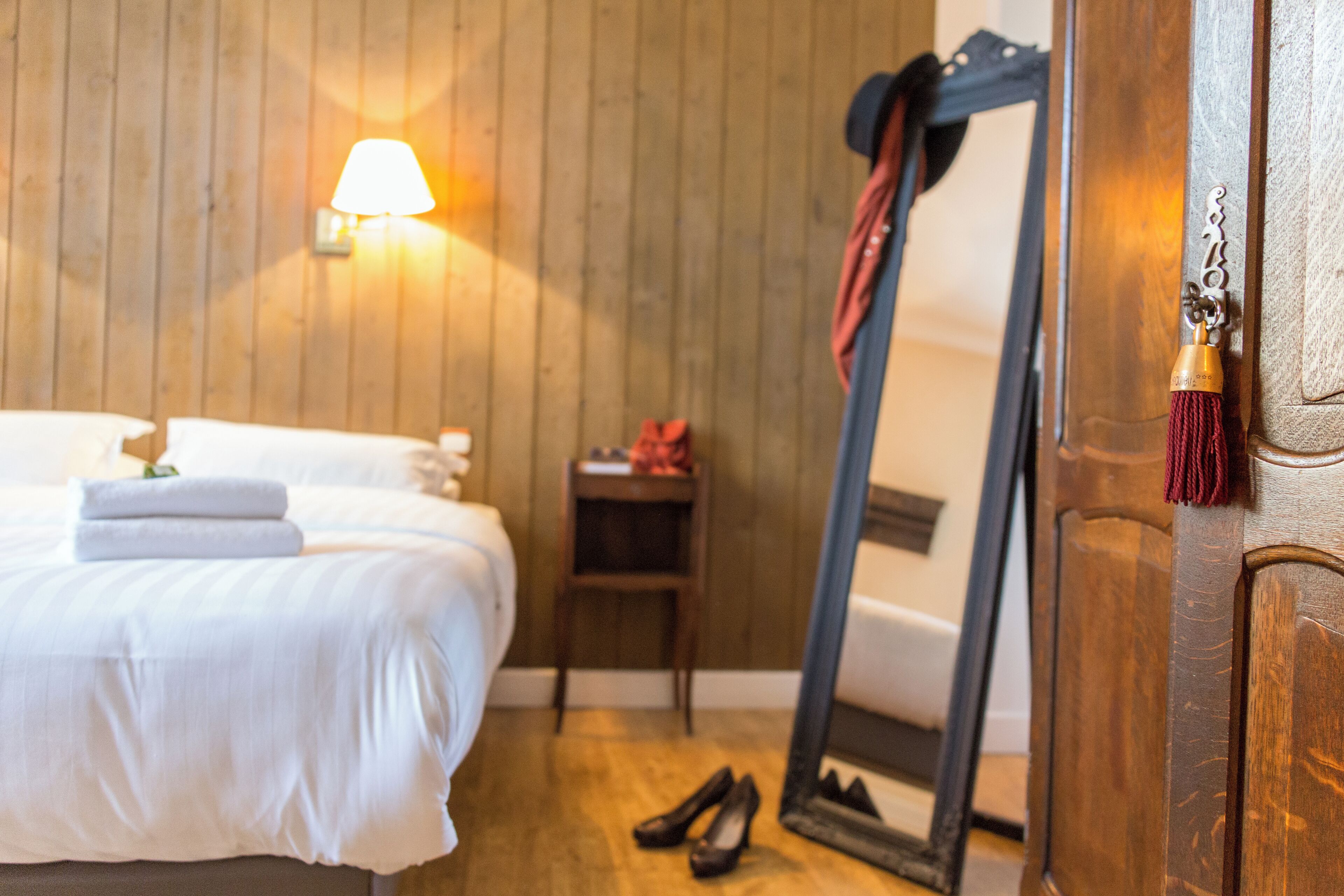 Un lit bien fait, avec une couette blanche et des serviettes pliées, un plancher en bois, une petite table de chevet en bois avec une lampe, et un miroir appuyé contre une porte en bois.