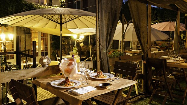 Een buitenterras met houten tafels en stoelen, parasols en tafels met kaarsen.