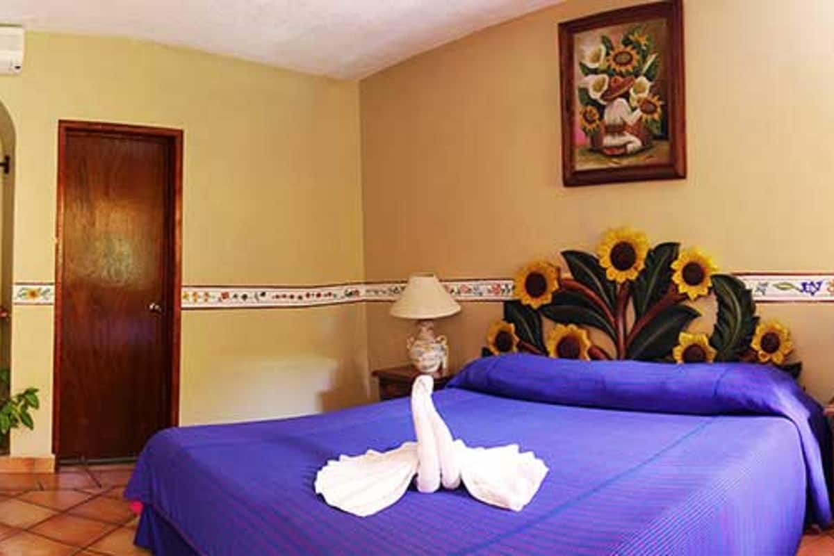 A hotel room with a bed, a wooden door, a painting, and a nightstand with a lamp.
