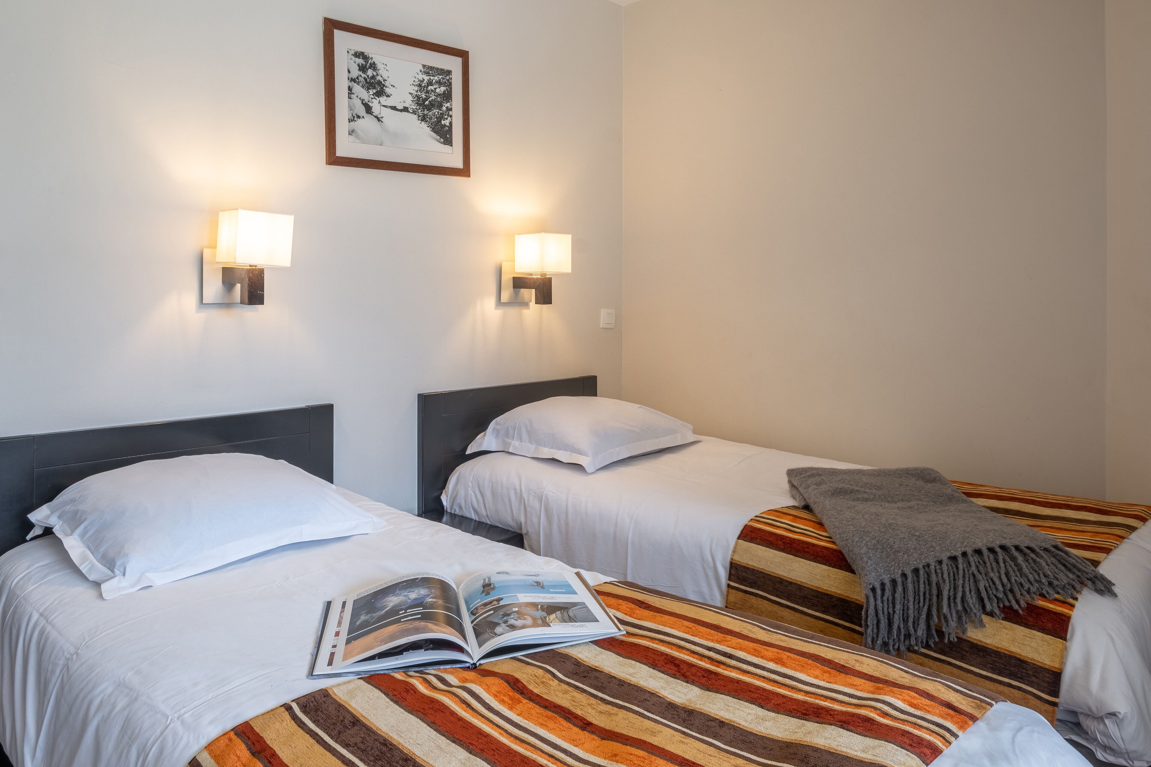 A hotel room with two beds, each with white linens and a striped blanket. There are two wall-mounted lamps on either side of the beds.