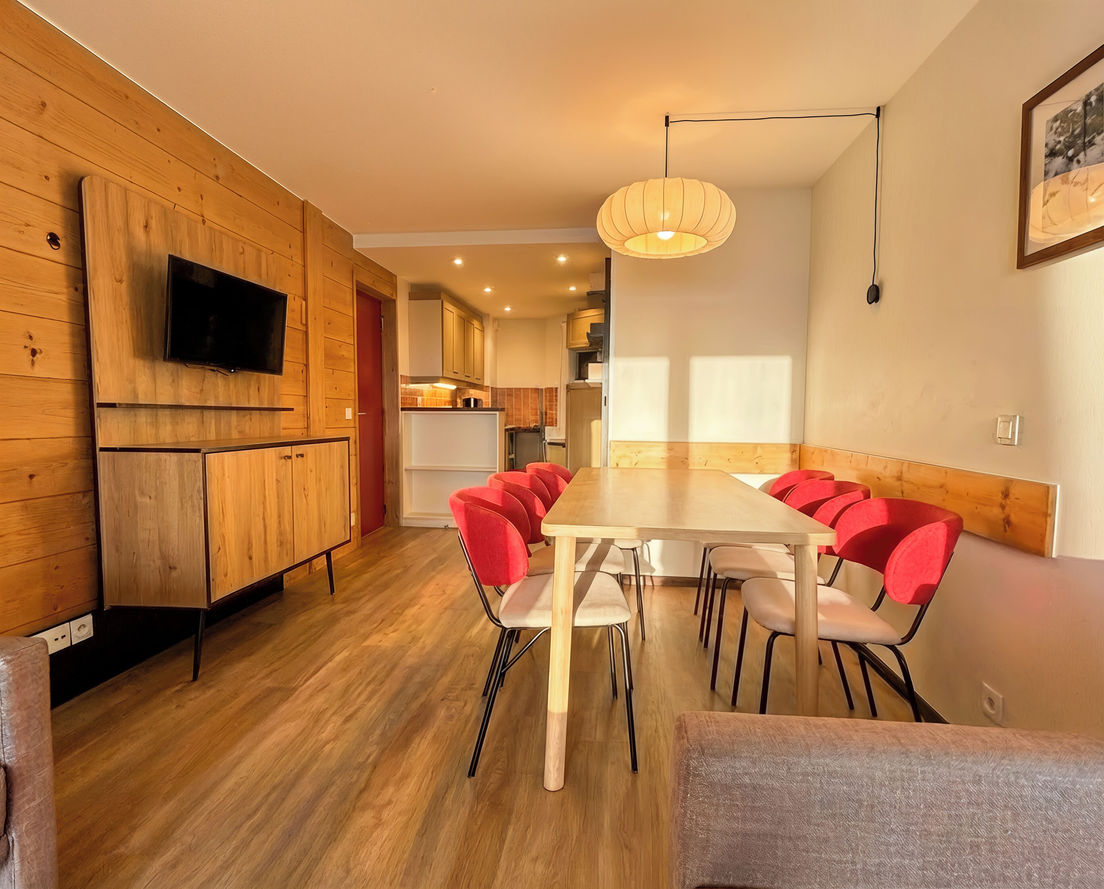 Eine moderne Küche mit Holzschränken, einem Flachbildfernseher und einem Essbereich mit einem Holztisch und roten Stühlen.