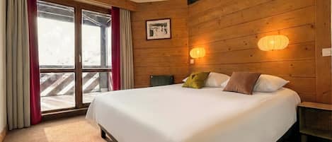 A bedroom with a large bed, wooden walls, a window with curtains, and two wall-mounted lights.