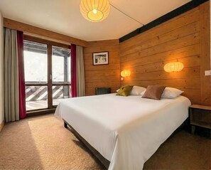 A bedroom with a large bed, wooden walls, a window with curtains, and two wall-mounted lights. - Pierre & Vacances Résidence le Tikal (Les Belleville)