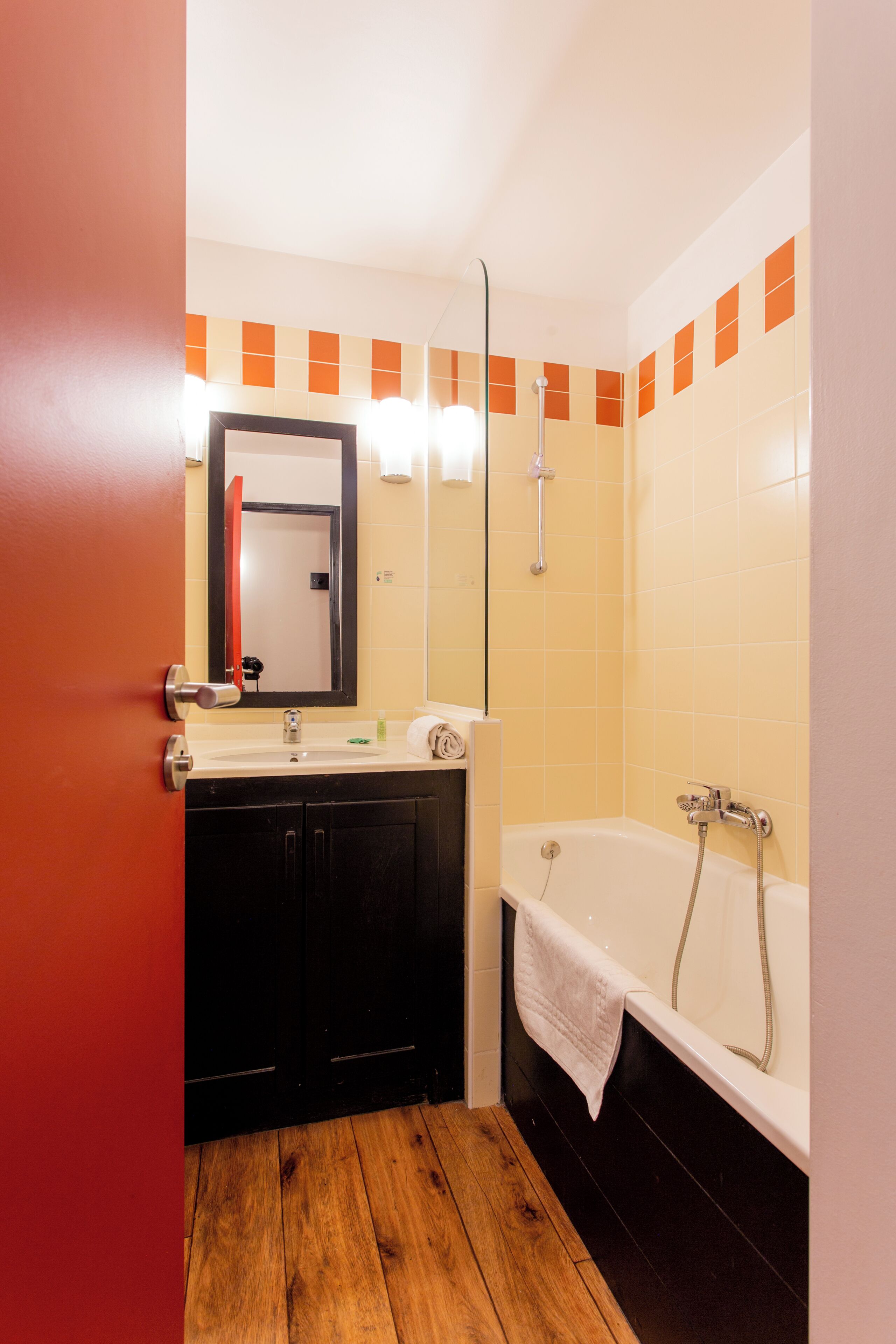 Une salle de bain avec un plancher en bois, un lavabo, un miroir, une baignoire et des murs recouverts de carreaux orange.