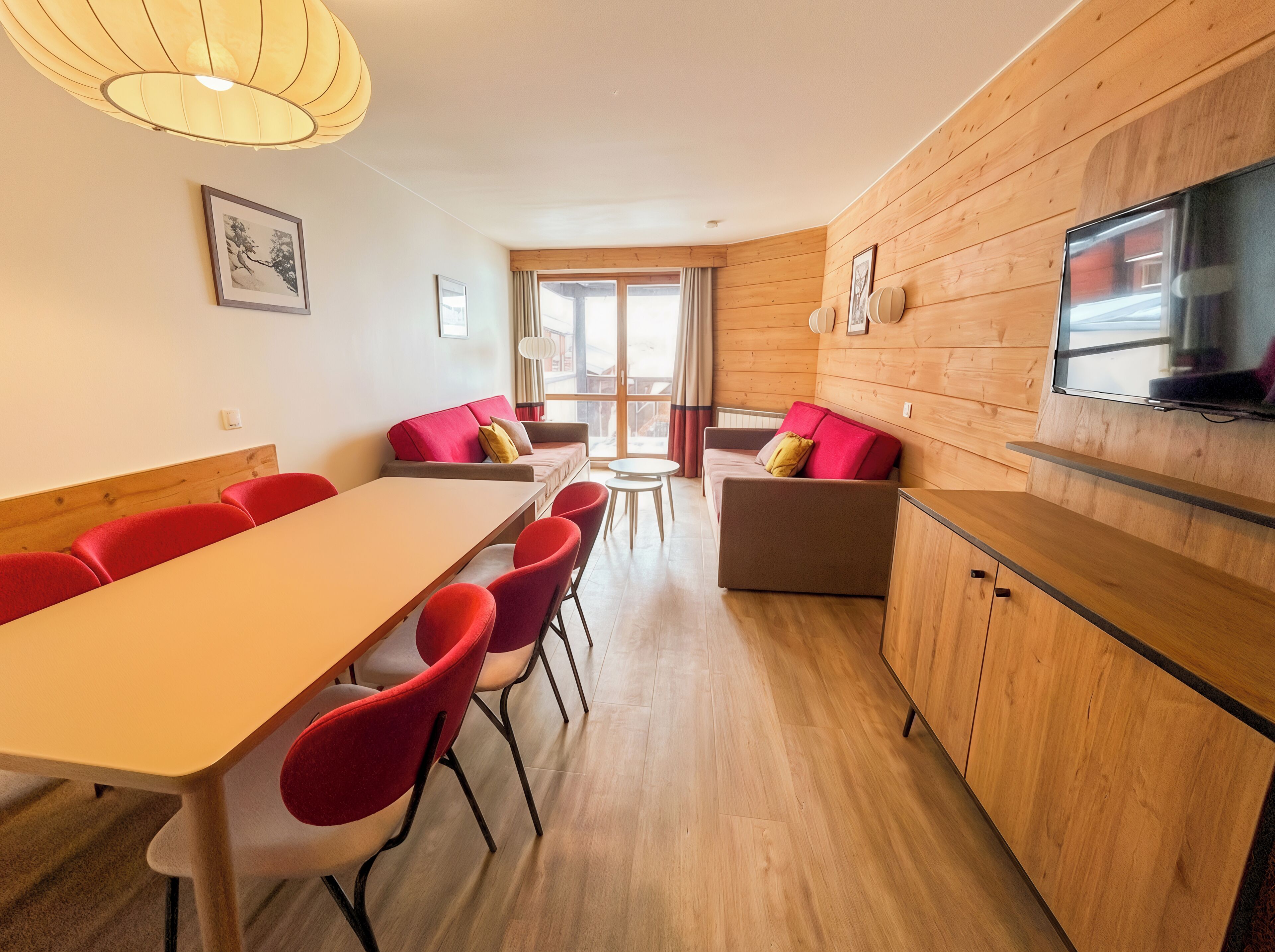 Ein modernes Zimmer mit Holzboden, einem langen Tisch mit roten Stühlen, einer Couch mit roten Kissen, einem Fernseher an der Wand und einem Fenster mit Vorhängen.