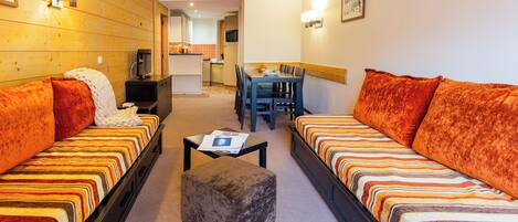 A hotel room with two sofas, a small ottoman, a dining table, and a kitchen area in the background.