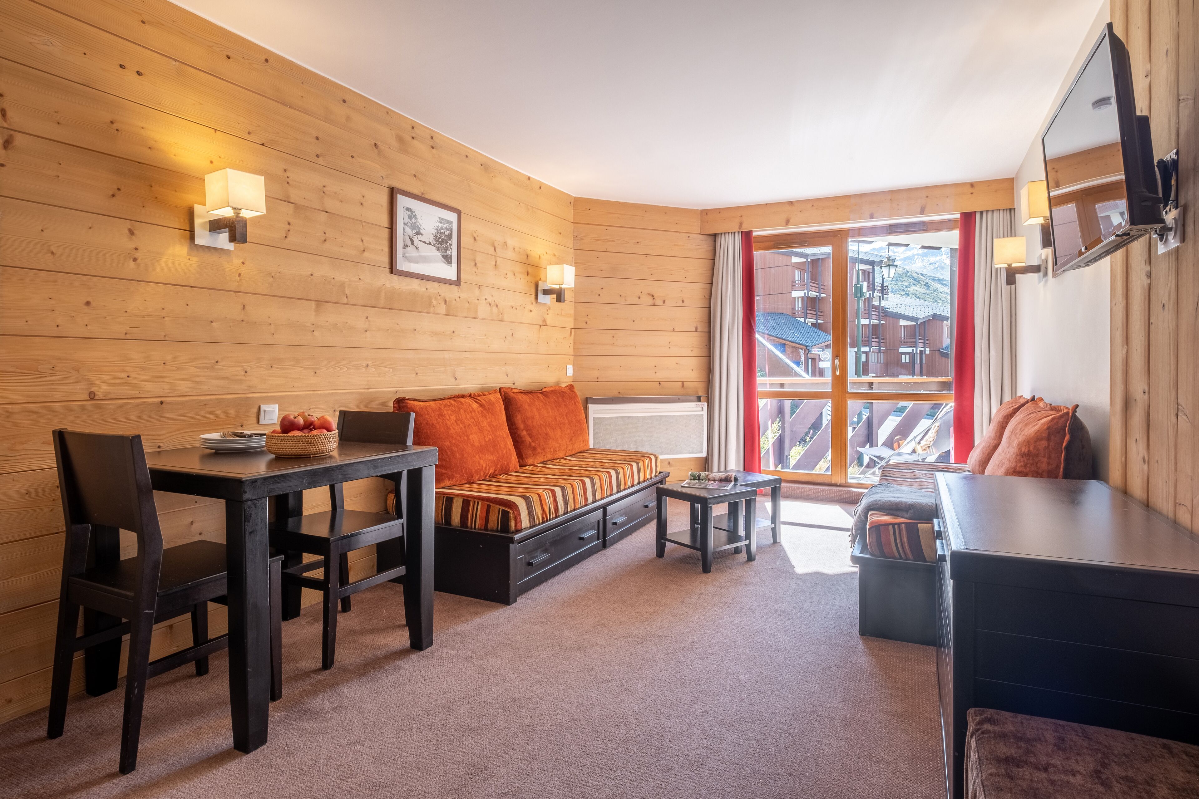 A cozy room with a wooden wall, a sofa, a dining table with chairs, a small table, and a TV mounted on the wall.