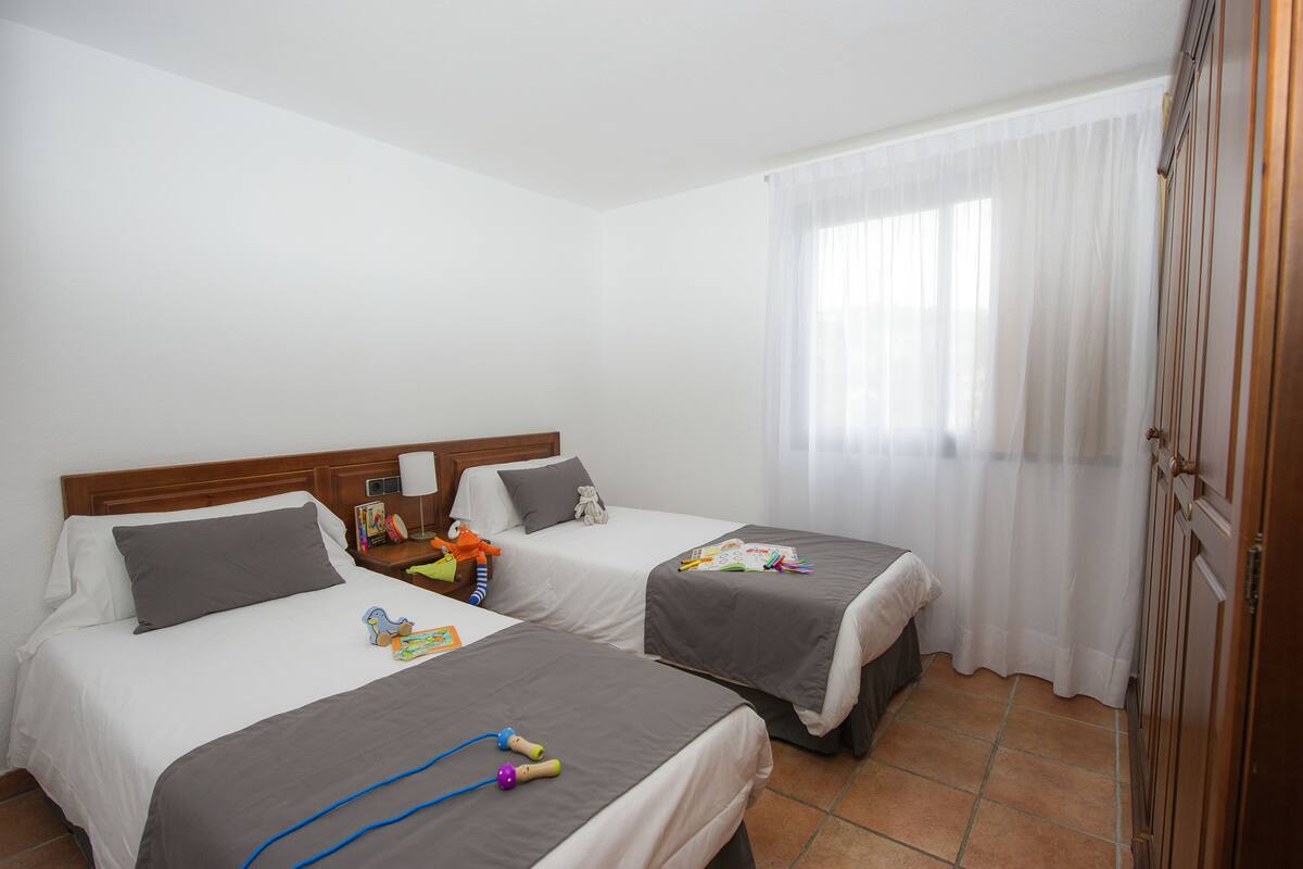 Habitación de hotel con dos camas, cabecera de madera, mesita de noche con lámpara y ventana con cortinas traslúcidas.