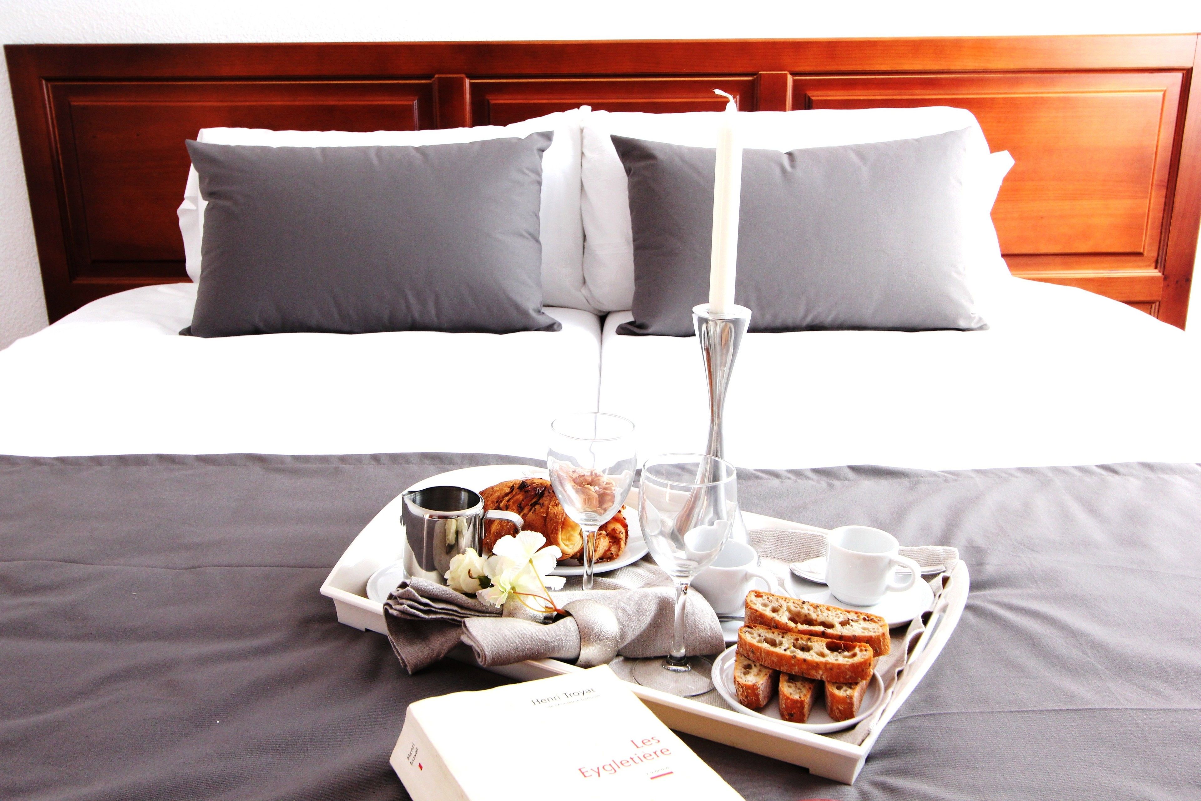 Una cama bien hecha con ropa de cama gris y un cabecero de madera, una bandeja con alimentos para el desayuno, incluyendo pan, un libro titulado 'Les Eygleterie', y una vela.