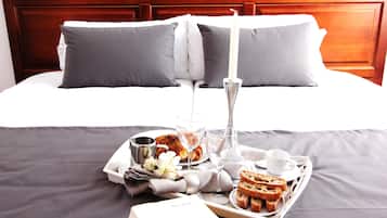 Una cama bien hecha con ropa de cama gris y un cabecero de madera, una bandeja con alimentos para el desayuno, incluyendo pan, un libro titulado 'Les Eygleterie', y una vela.