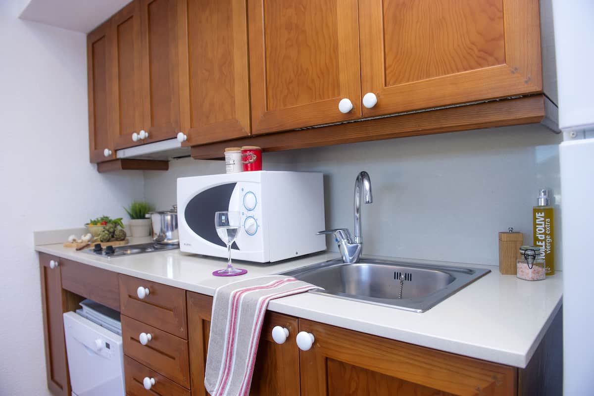 Una cocina compacta con armarios de madera, una encimera blanca, microondas, fregadero y escurreplatos.