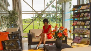 A woman seated on a wicker sofa with a laptop, surrounded by a glass wall with a view of a green landscape.