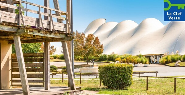 A modern building with a unique, curved roof structure and a wooden pergola in the foreground. - Pierre & Vacances Résidence Les Rives de la Seugne (Jonzac)