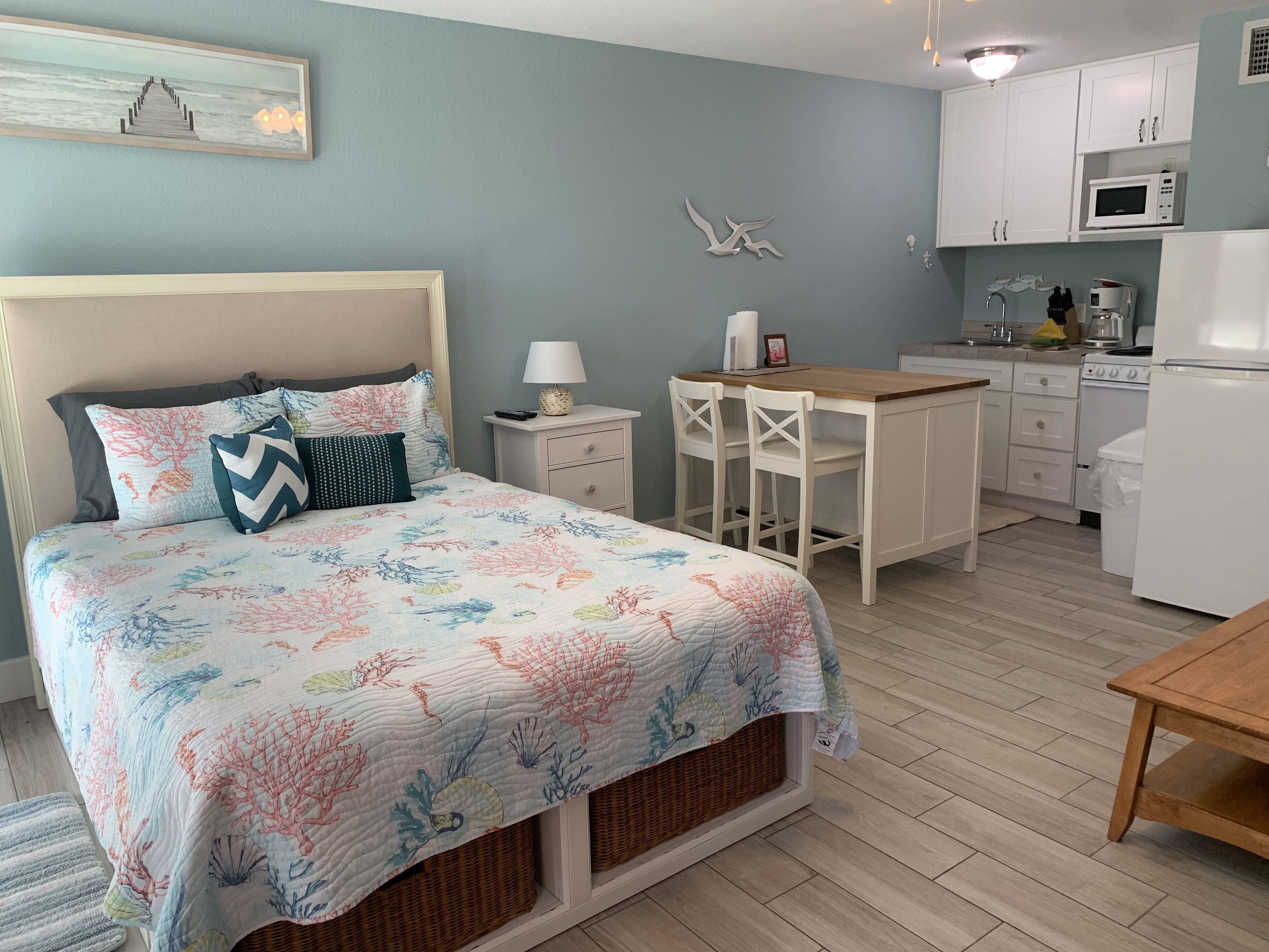 A bedroom with a bed, bedside table, lamp, and a small kitchenette with a microwave, stove, and refrigerator.