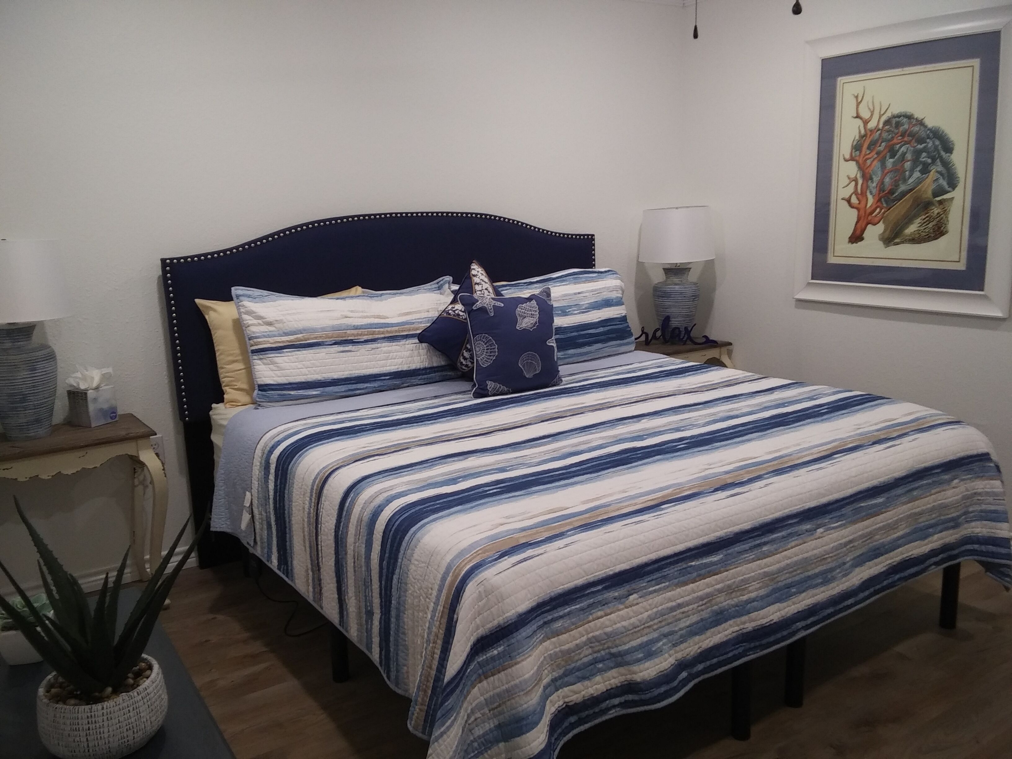 Ein Schlafzimmer mit Bett, Kissen, Nachttisch, Lampe, Wandbild und Blumentopf.