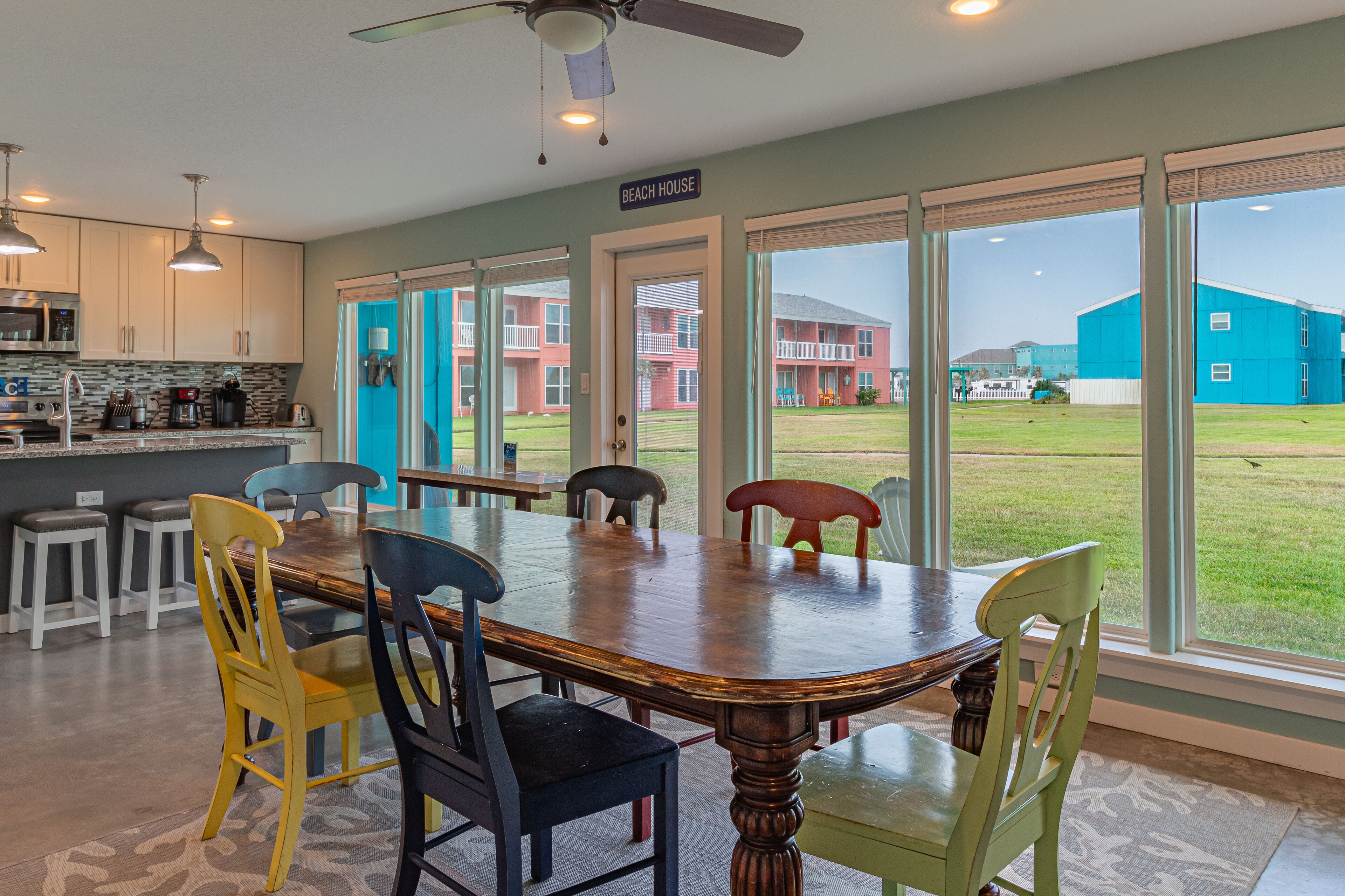 A modern kitchen with stainless steel appliances, a dining area with a wooden table and colorful chairs, and large windows offering a view of a building and grassy area.
