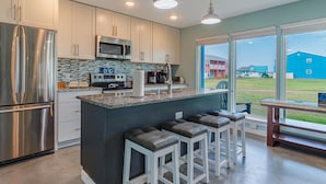 A modern kitchen with stainless steel appliances, a large island with bar stools, and a view of a blue building outside. - Executive Keys Condominiums on the Beach (Port Aransas)