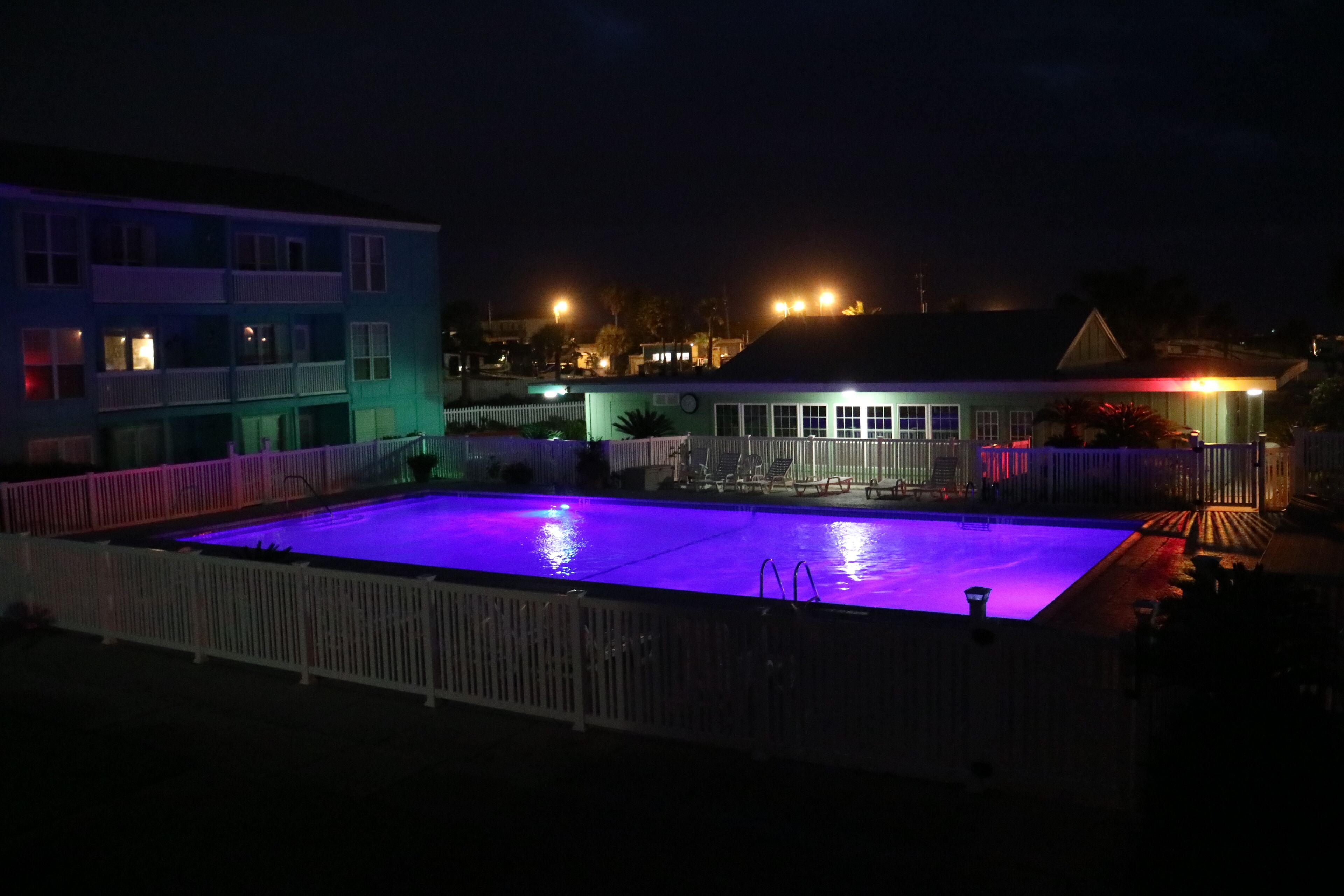 Une piscine entourée d’une clôture blanche, elle-même bordée de bâtiments dont les fenêtres sont illuminées.