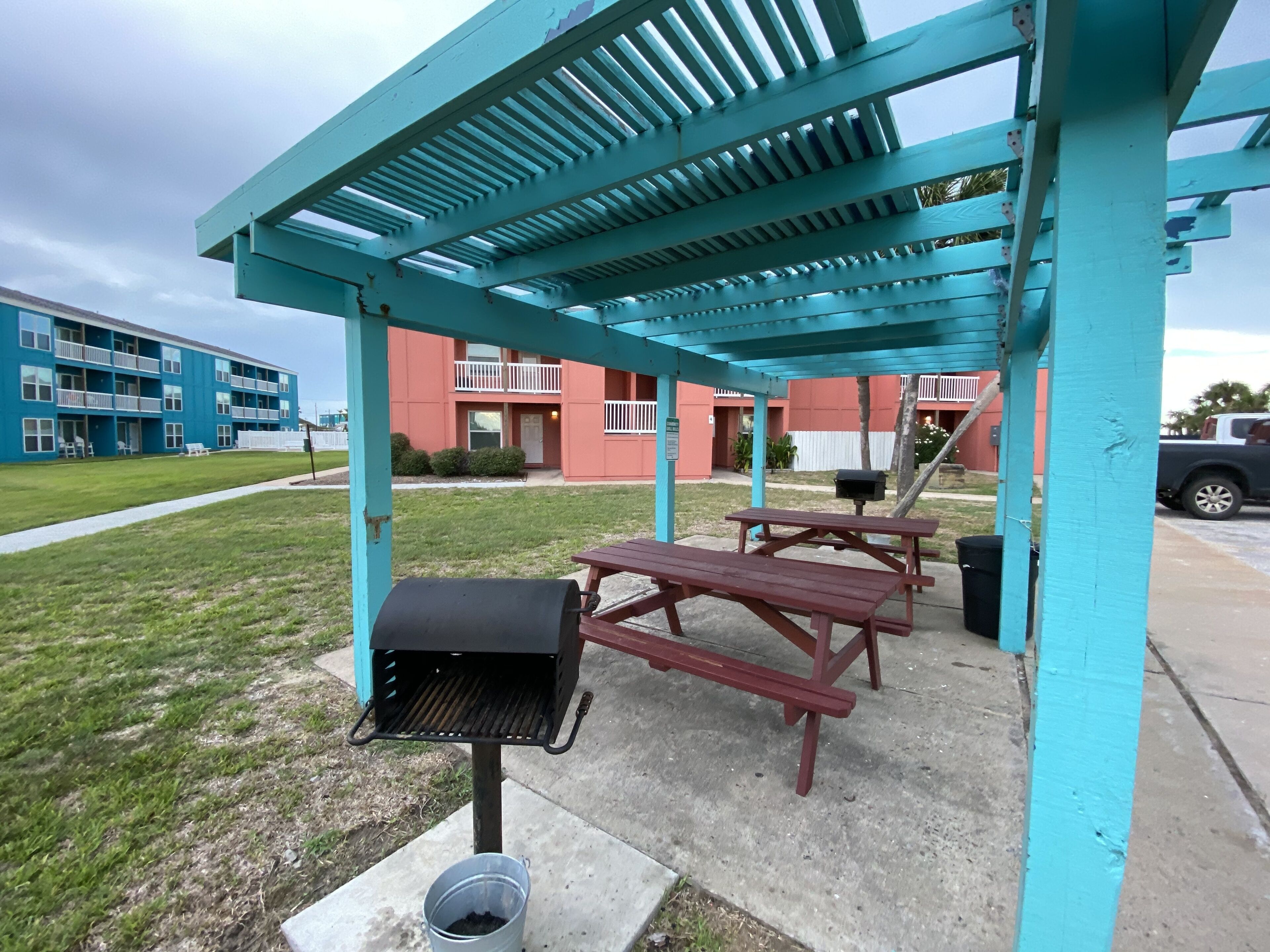 Un patio exterior con una parrilla, mesas de picnic y una zona cubierta con techo azul.