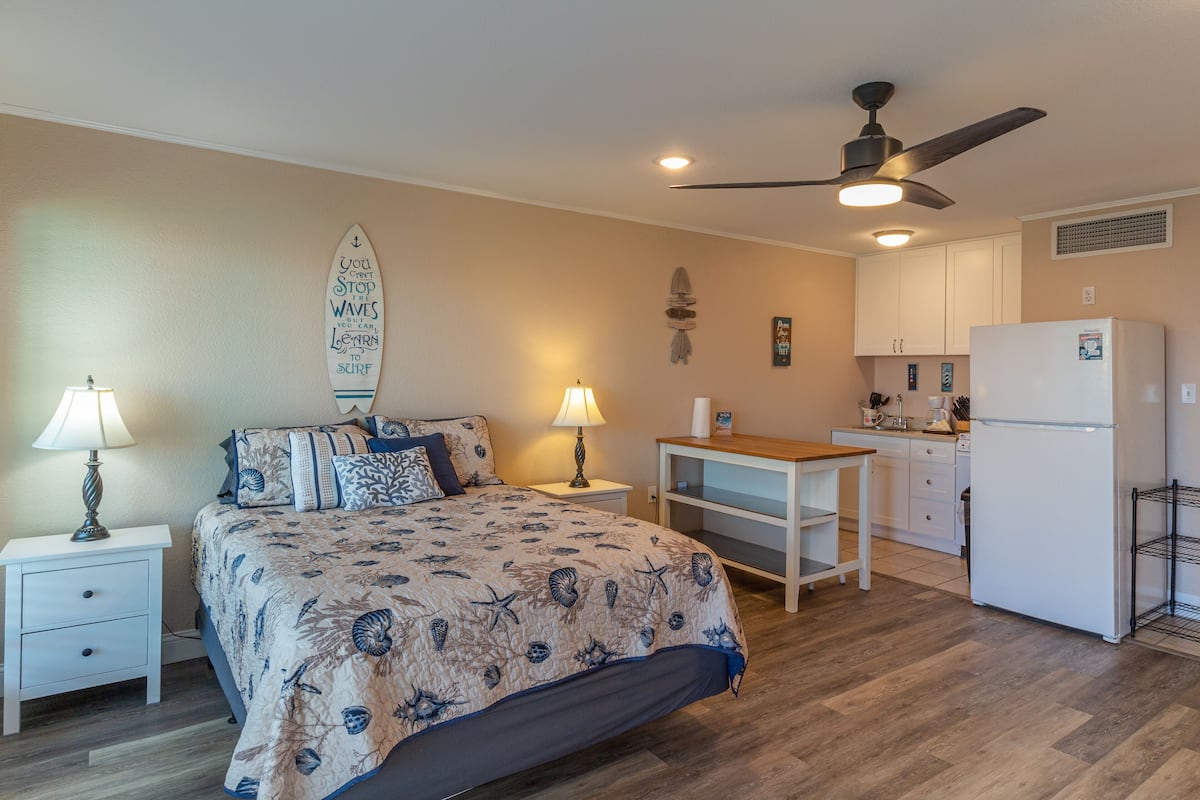 A bedroom with a bed, bedside tables, a desk, a refrigerator, and a ceiling fan.