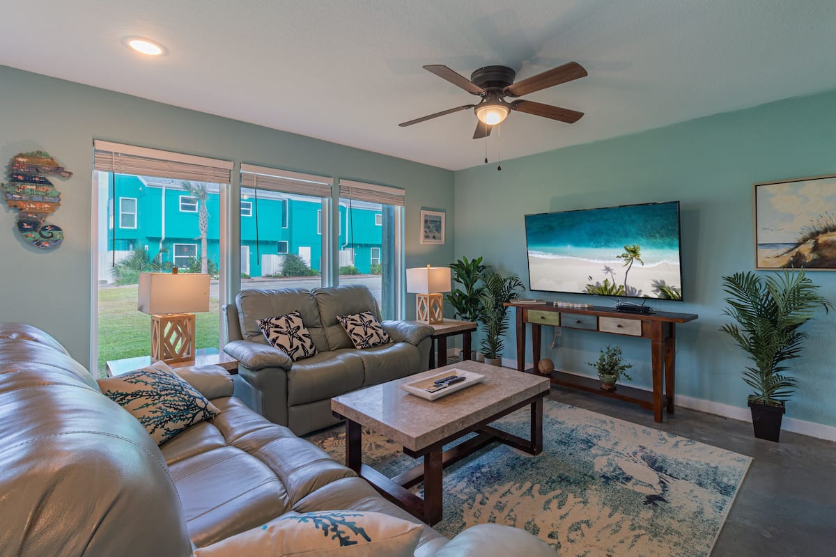 A living room with a sofa, coffee table, and TV. There are windows, a ceiling fan, and a painting on the wall.
