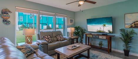 A living room with a sofa, coffee table, and TV. There are windows, a ceiling fan, and a painting on the wall.