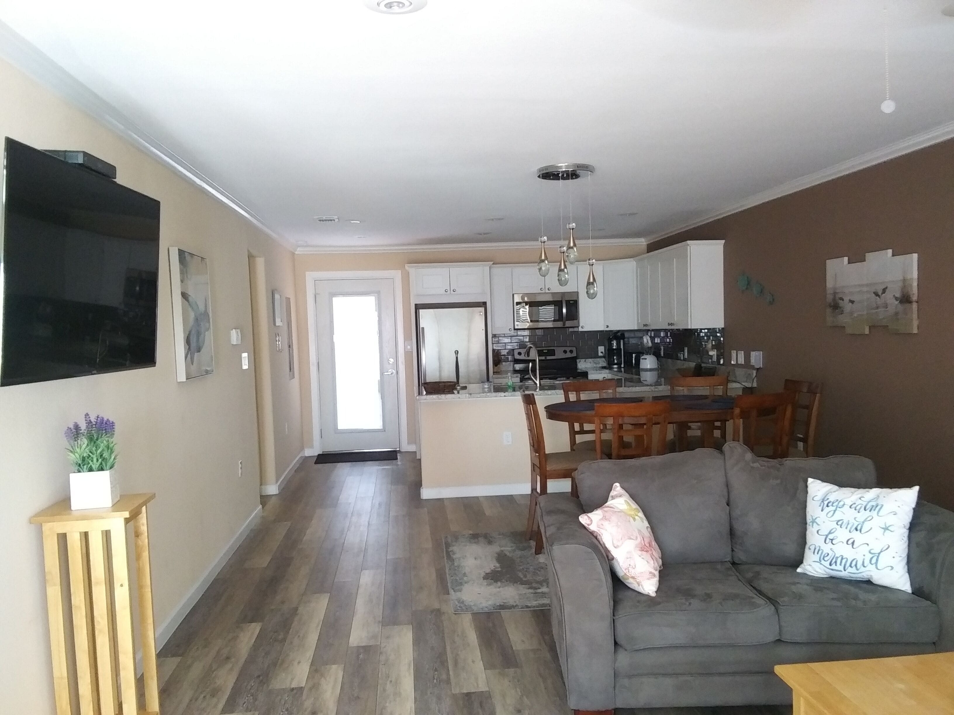 A modern living room with a grey sofa, wooden coffee table, and a dining area with a wooden table and chairs. A kitchen with white cabinets and stainless steel appliances is visible in the background.
