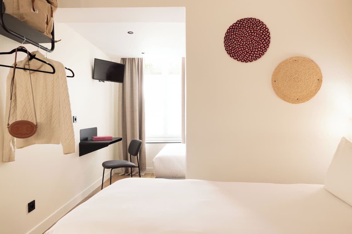 A hotel room with a bed, a chair, a TV mounted on the wall, a shelf with a book, and a window with curtains.