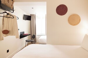 Habitación de hotel con cama, una silla, televisor montado en la pared, estante con un libro y ventana con cortinas.