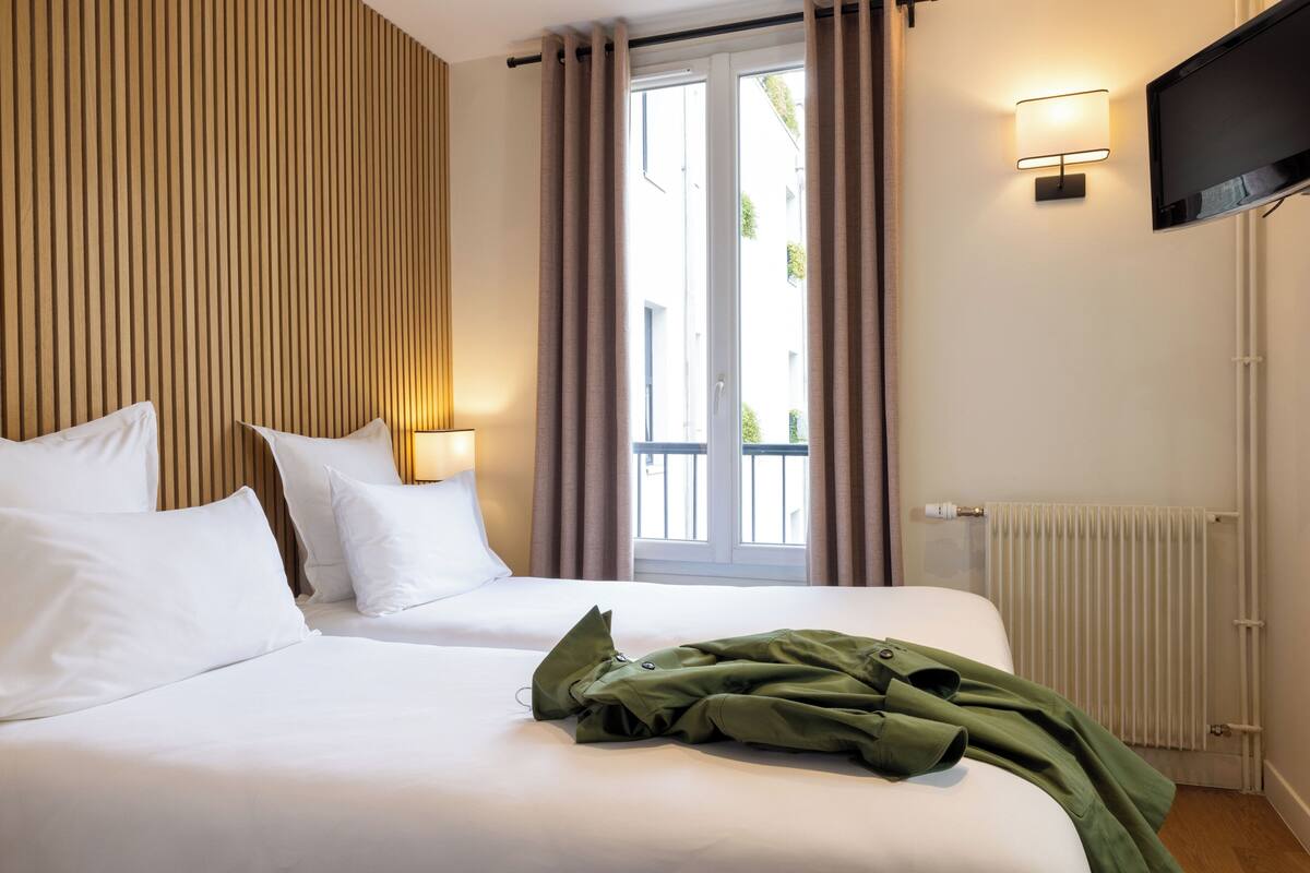 A hotel room with a bed, a window with curtains, a wall-mounted TV, and a radiator.