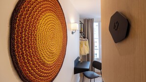 A room with a large, circular, textured wall art piece, a wall clock, a small desk with a chair, and a window with curtains. - Korner Montmartre (Paris)