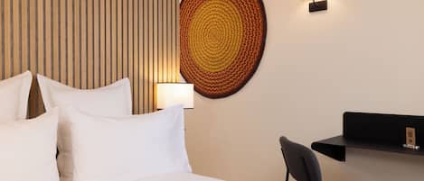 A hotel room with a bed, pillows, a wall-mounted lamp, a round woven wall art, and a small desk with a chair.
