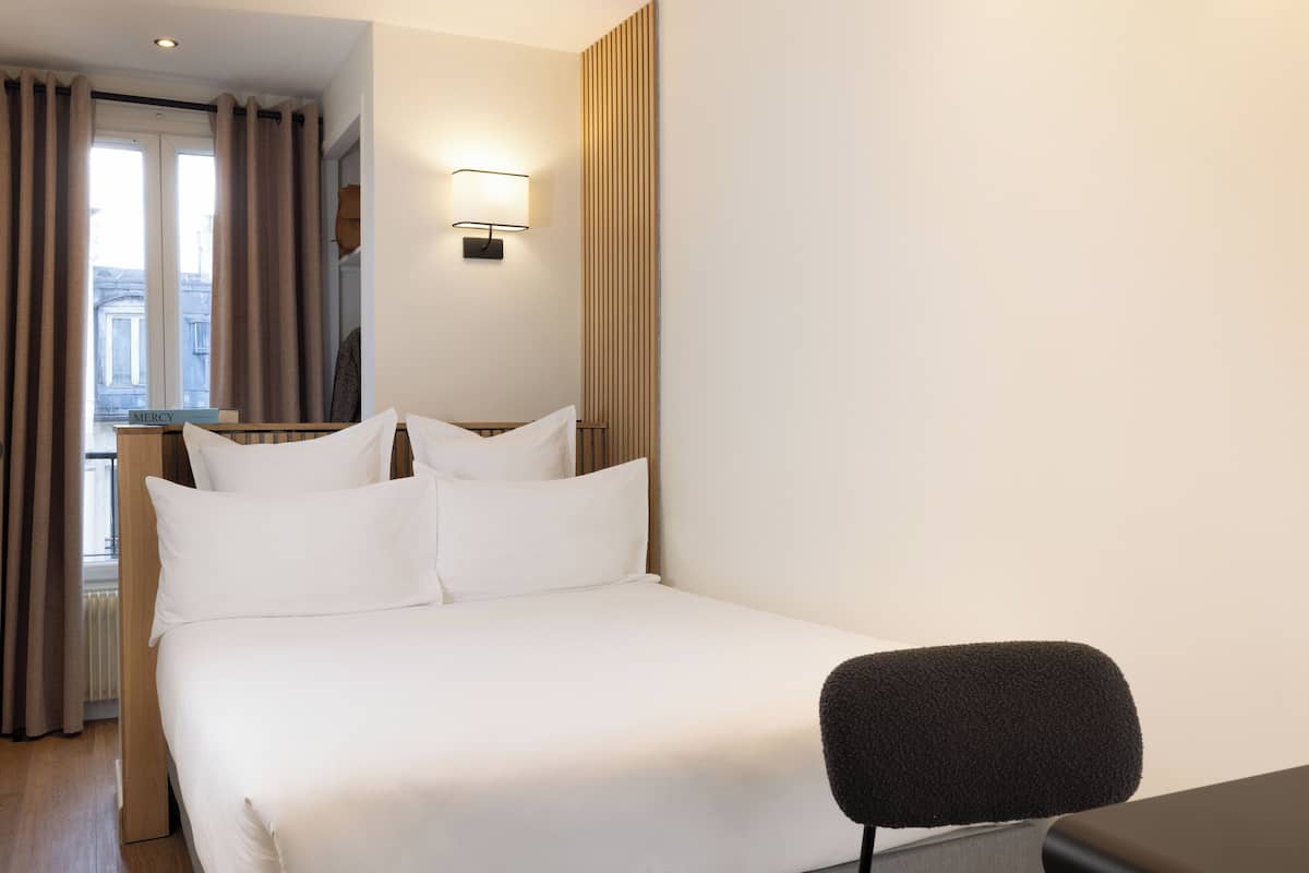 A hotel room with a bed, a desk, a chair, a lamp, and a window with curtains.