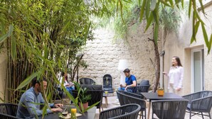 An outdoor patio area with wicker chairs and tables, a stone wall, and greenery. - Korner Montmartre (Paris)