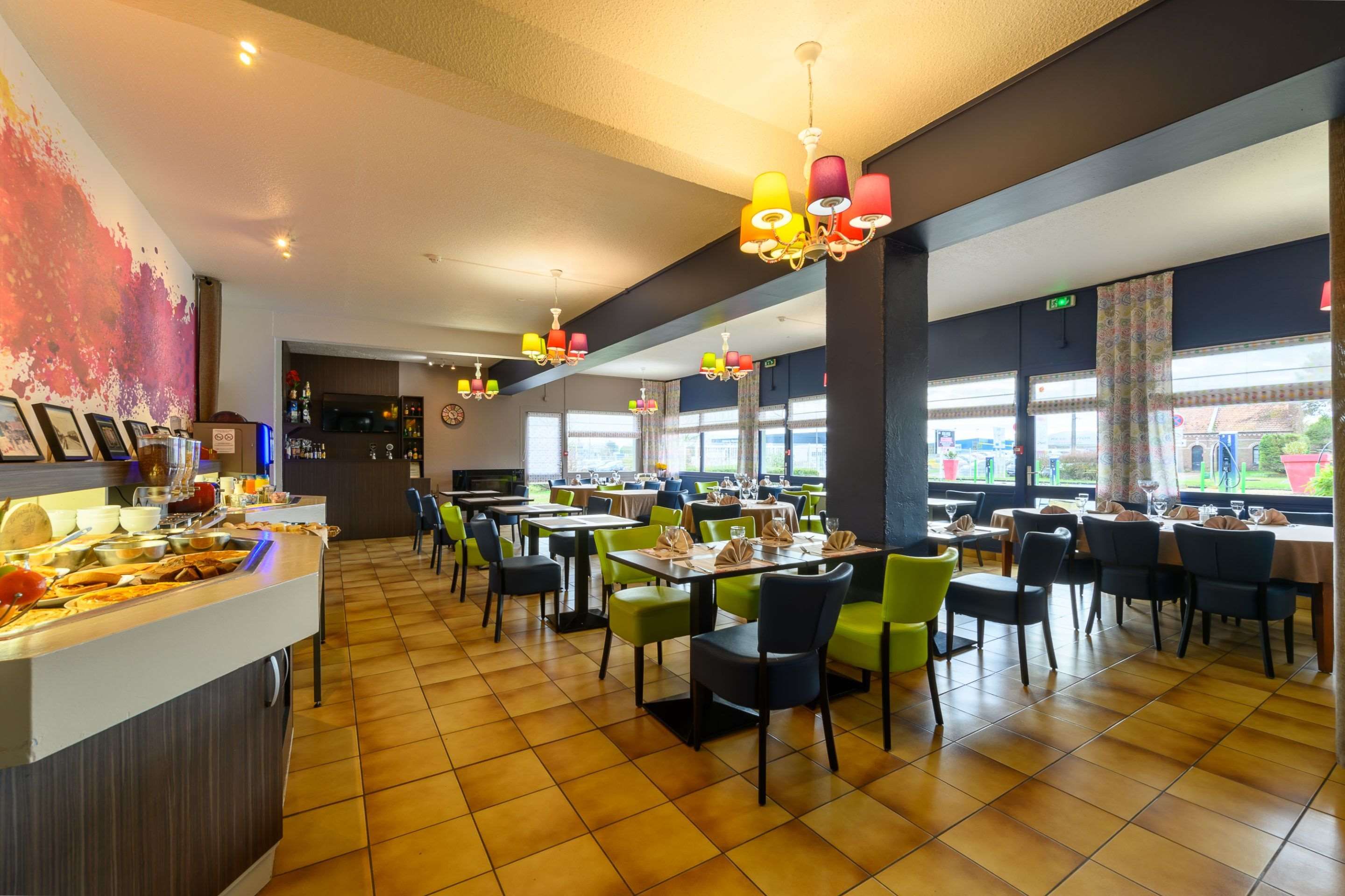 Ein Restaurant mit mehreren Tischen und Stühlen, einer Theke mit Speisen sowie Blick ins Freie durch große Fenster.