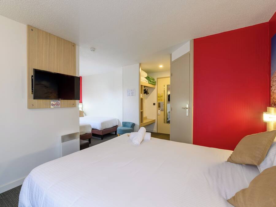 A hotel room with a large bed, a TV mounted on the wall, a desk with a chair, and a red accent wall.