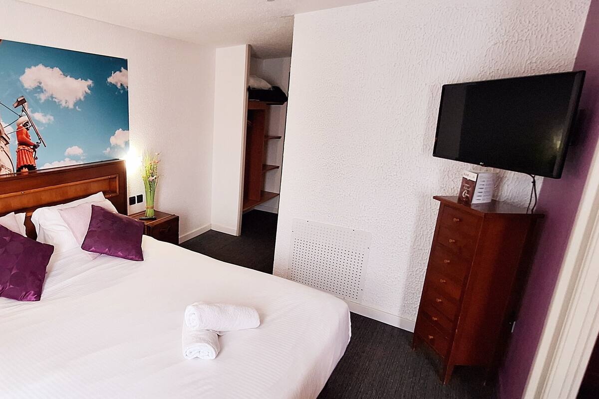 Habitación de hotel con una cama grande, dos almohadas moradas, una toalla blanca, un cuadro de un edificio histórico, un televisor y un aparador de madera.