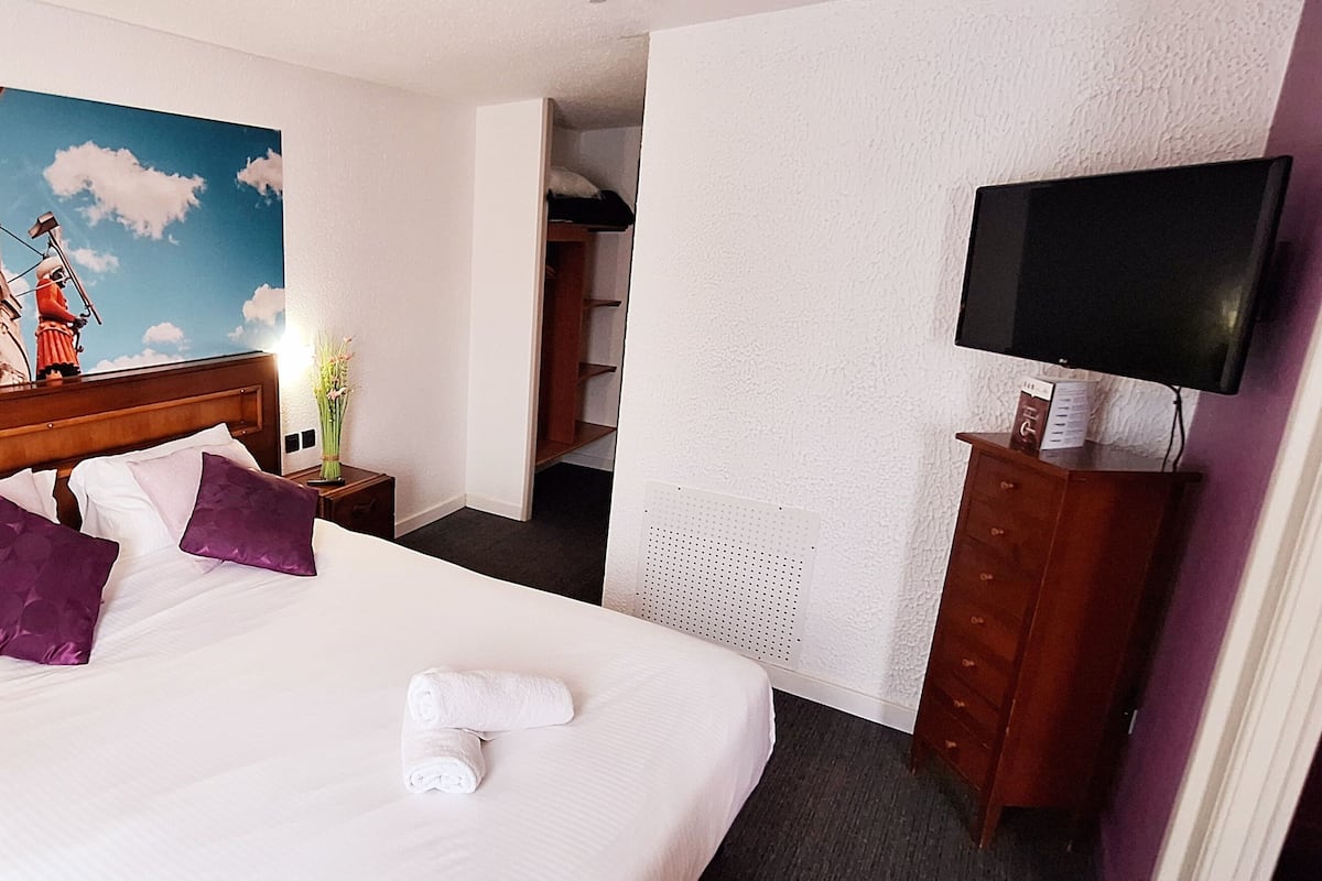 A hotel room with a large bed, two purple pillows, a white towel, a painting of a historical building, a television, and a wooden dresser.