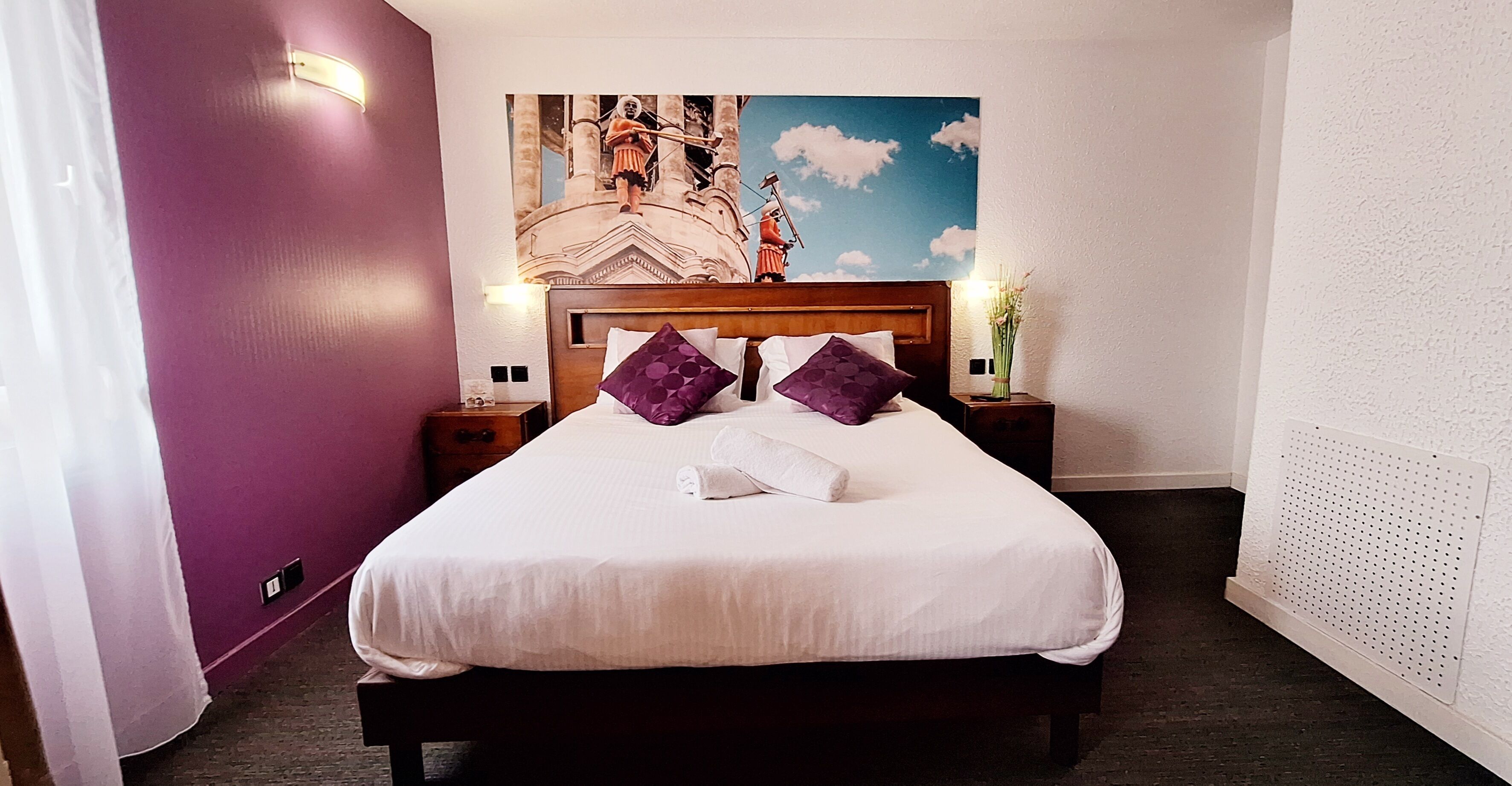 Una habitación de hotel con cama, almohadones morados, una mesita de noche, una lámpara de pared y un adorno mural.