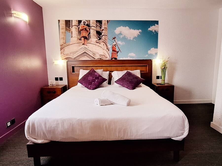 Una habitación de hotel con cama, almohadones morados, una mesita de noche, una lámpara de pared y un adorno mural.