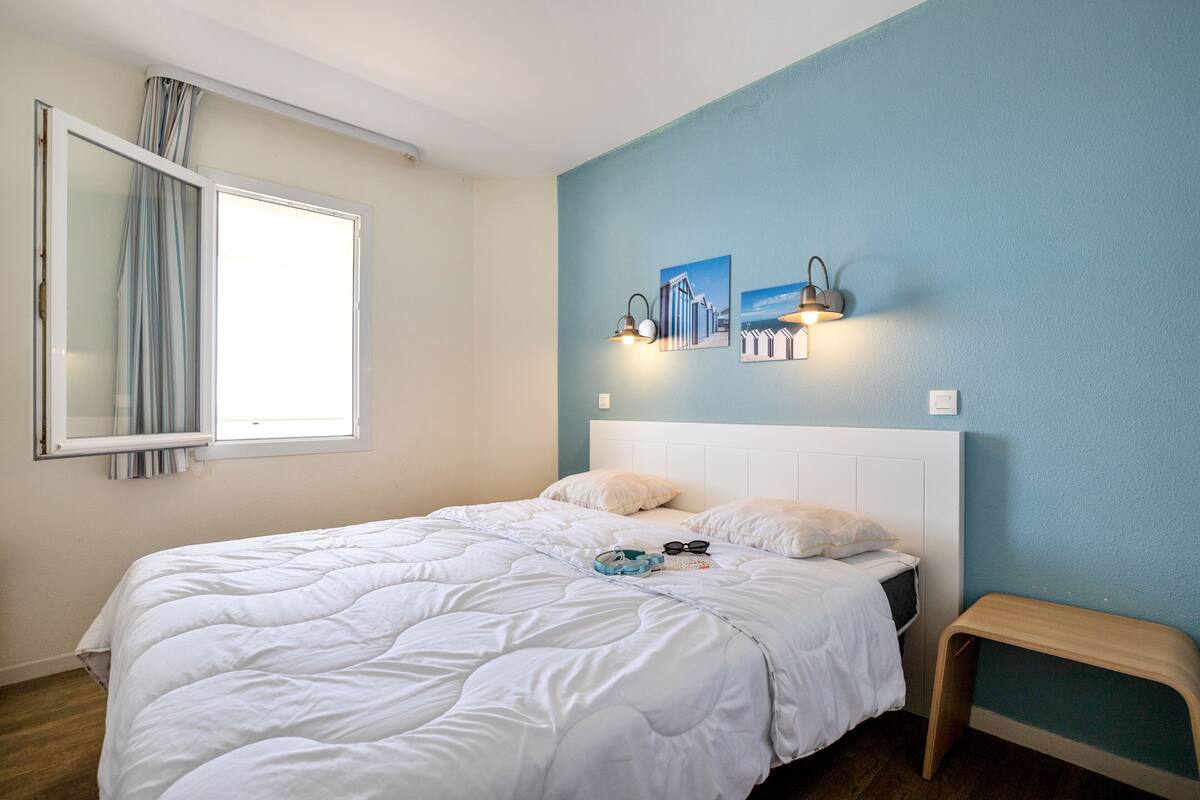 Un dormitorio con cama, mesita de noche, ventana con cortinas y una obra de arte en la pared.