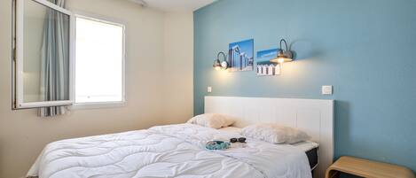 Un dormitorio con cama, mesita de noche, ventana con cortinas y una obra de arte en la pared.