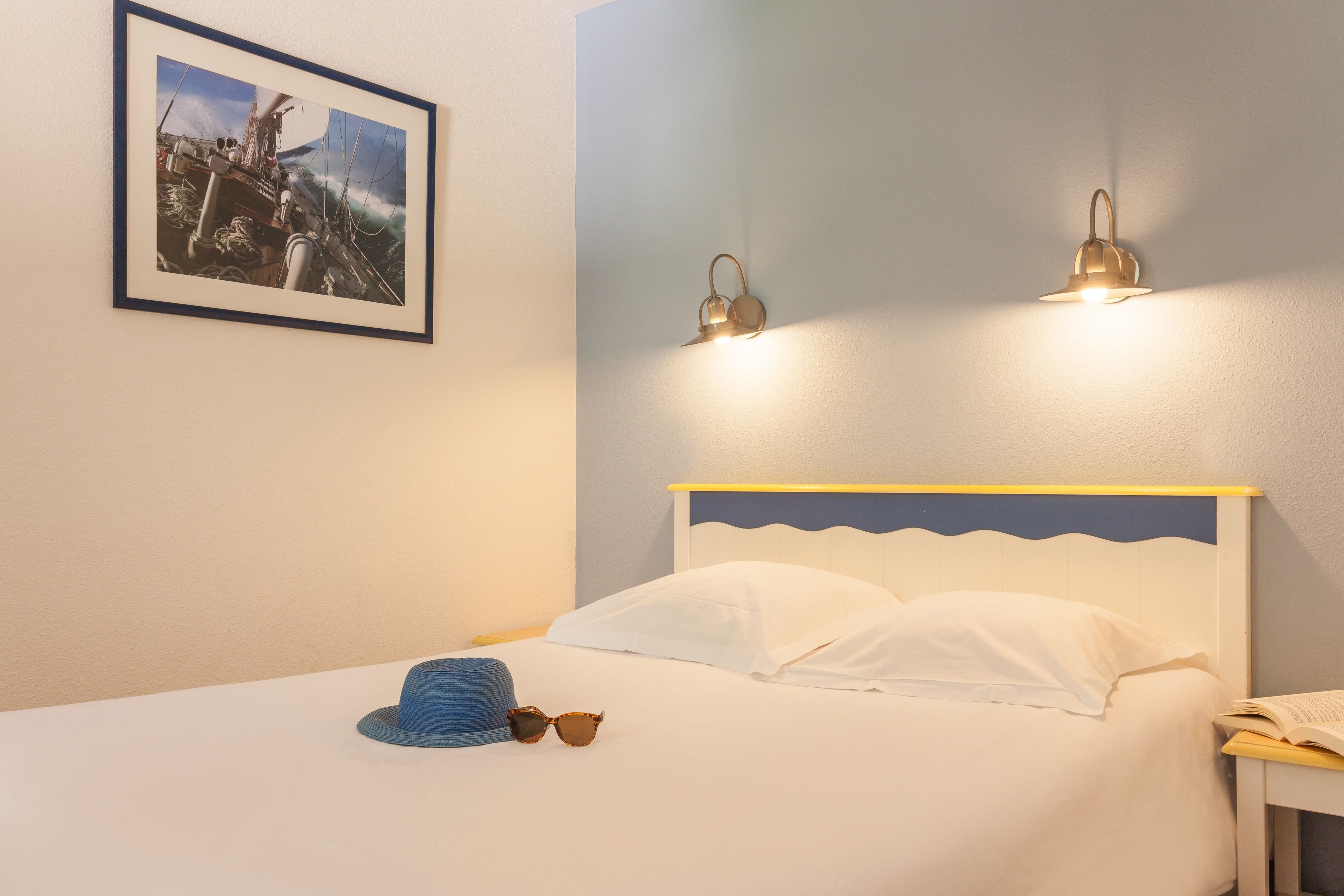 Un lit bien fait, avec du linge de lit blanc, un chapeau bleu et des lunettes de soleil posés sur le lit. Un tableau encadré représentant un navire est accroché au mur. Deux lampes fixées au mur assurent l’éclairage.
