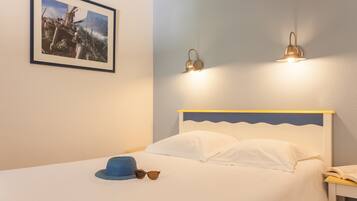 Un lit bien fait, avec du linge de lit blanc, un chapeau bleu et des lunettes de soleil posés sur le lit. Un tableau encadré représentant un navire est accroché au mur. Deux lampes fixées au mur assurent l’éclairage.