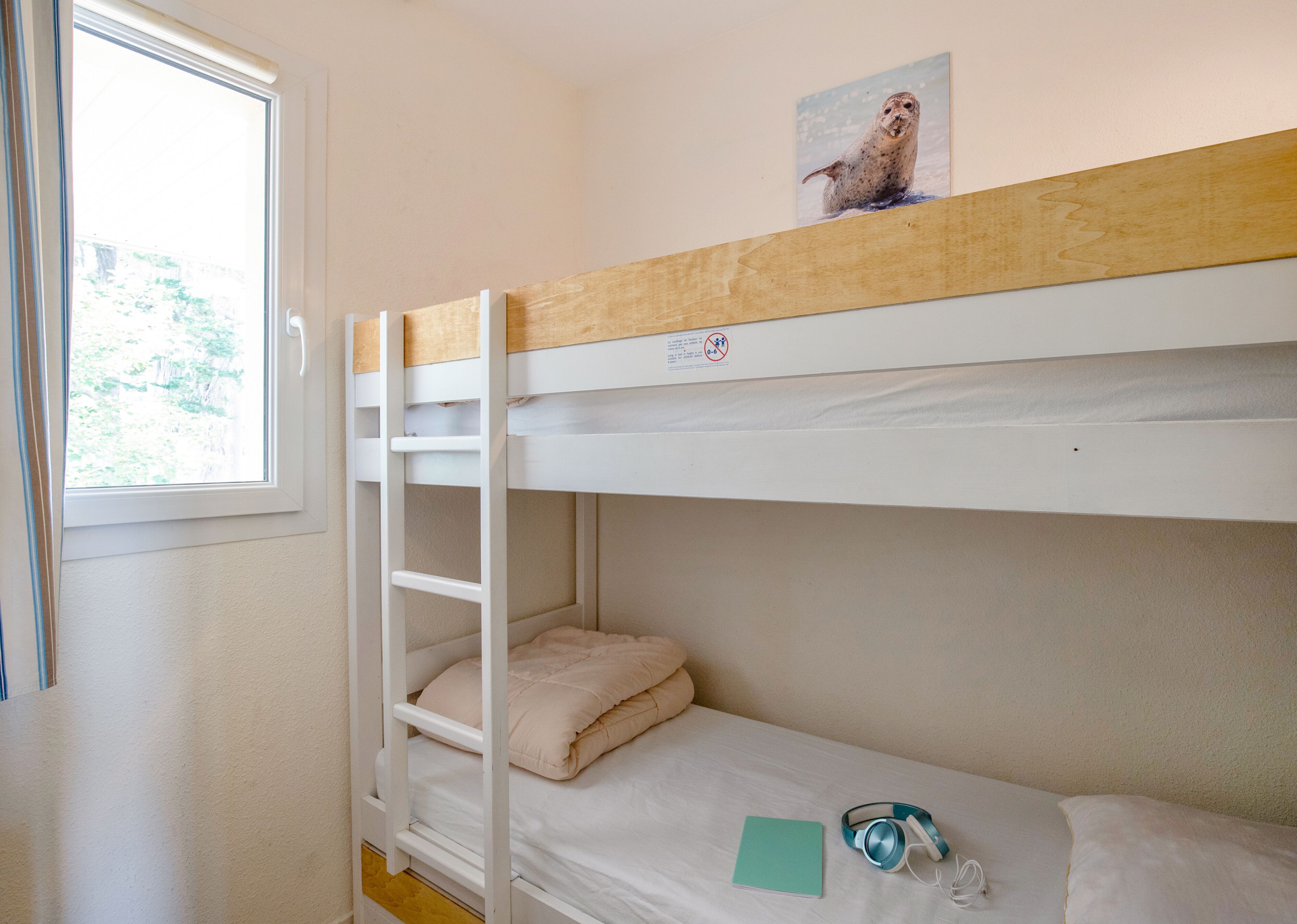 Une chambre avec un lit superposé, une fenêtre, une image accrochée au lit supérieur et une paire d’écouteurs posée sur le lit inférieur.