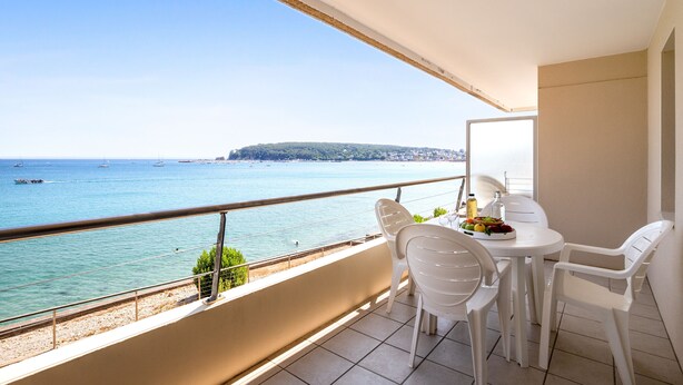 Un balcón con vistas al mar, sillas blancas y una mesa con frutero.