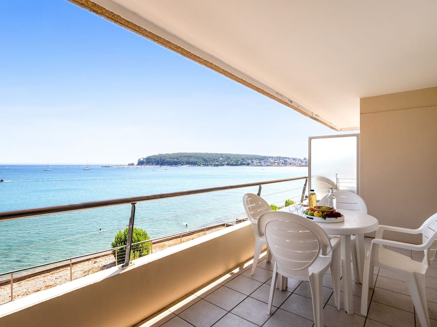 Un balcon avec vue sur la mer, des chaises blanches et une table sur laquelle se trouve un panier de fruits.