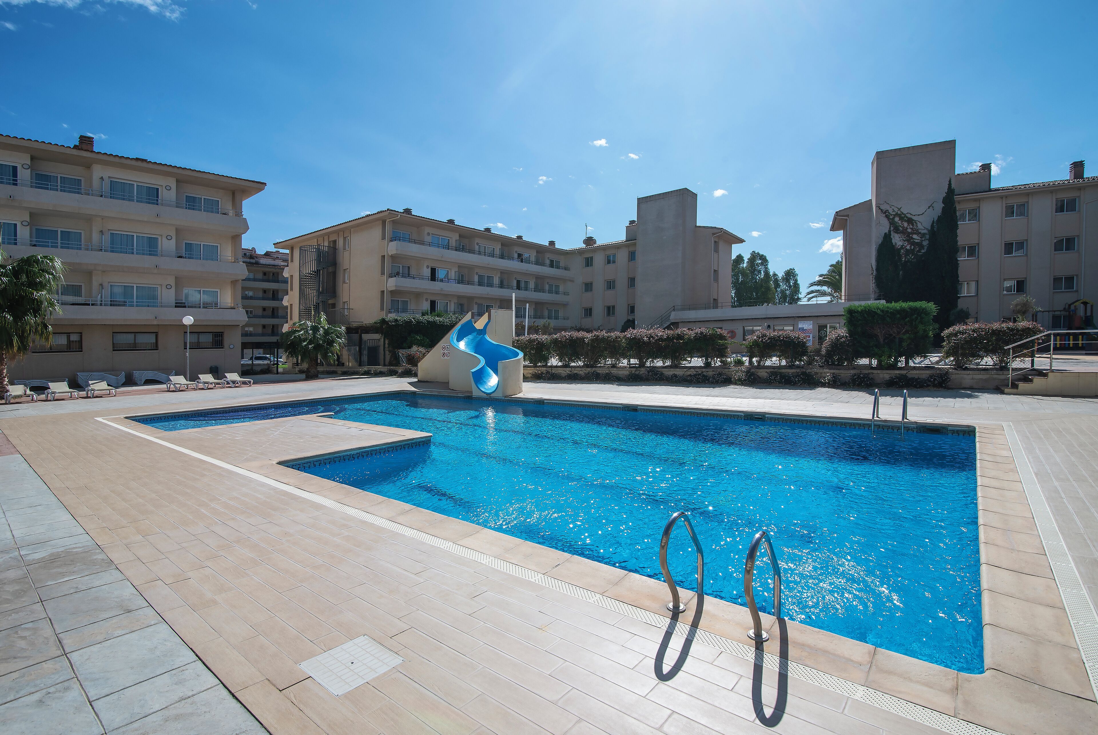 A swimming pool with a slide, surrounded by a tiled deck and modern buildings.