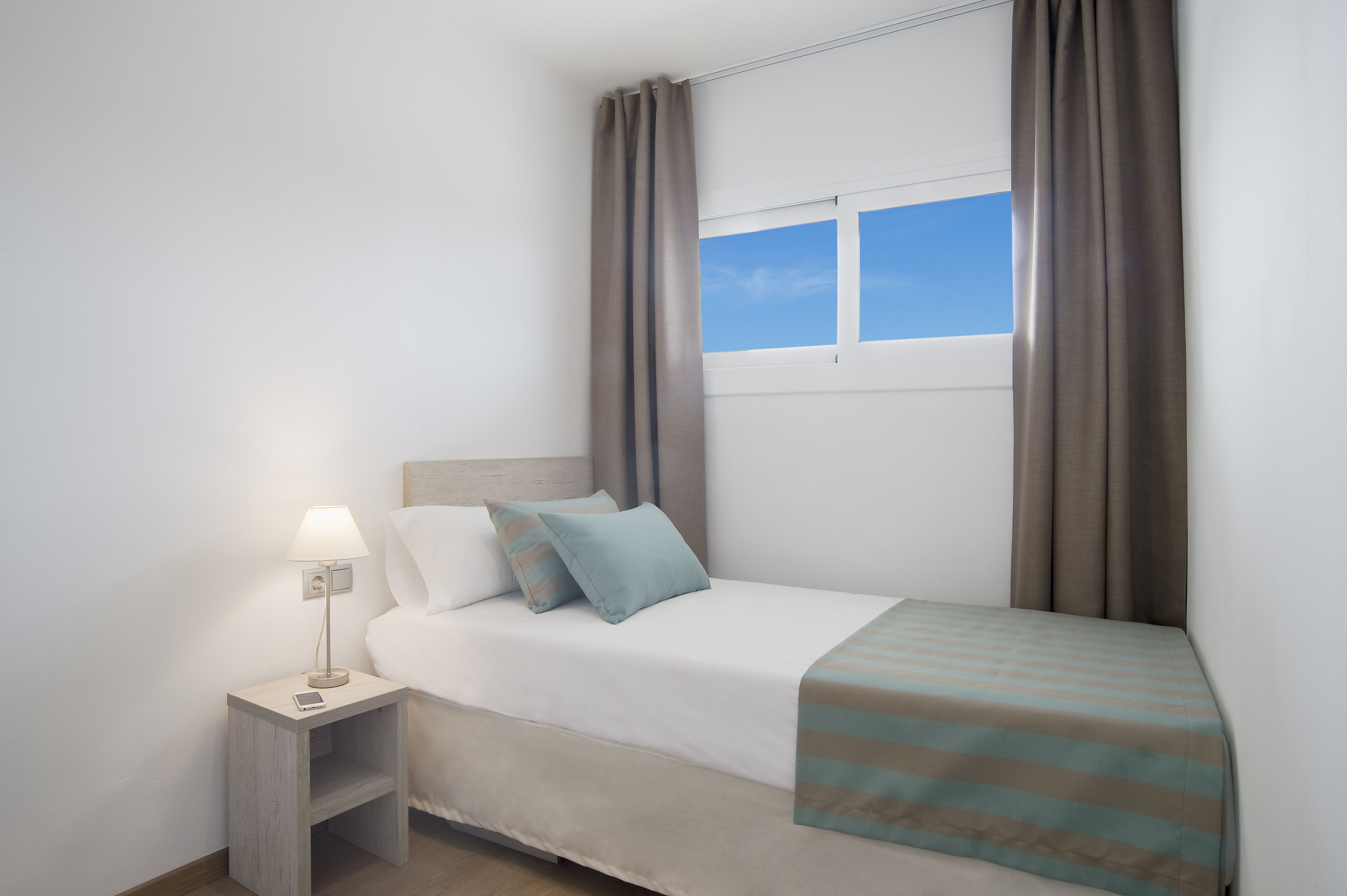 Una cama bien hecha con una manta a rayas, una lámpara de noche y un teléfono inteligente sobre una mesita de madera.